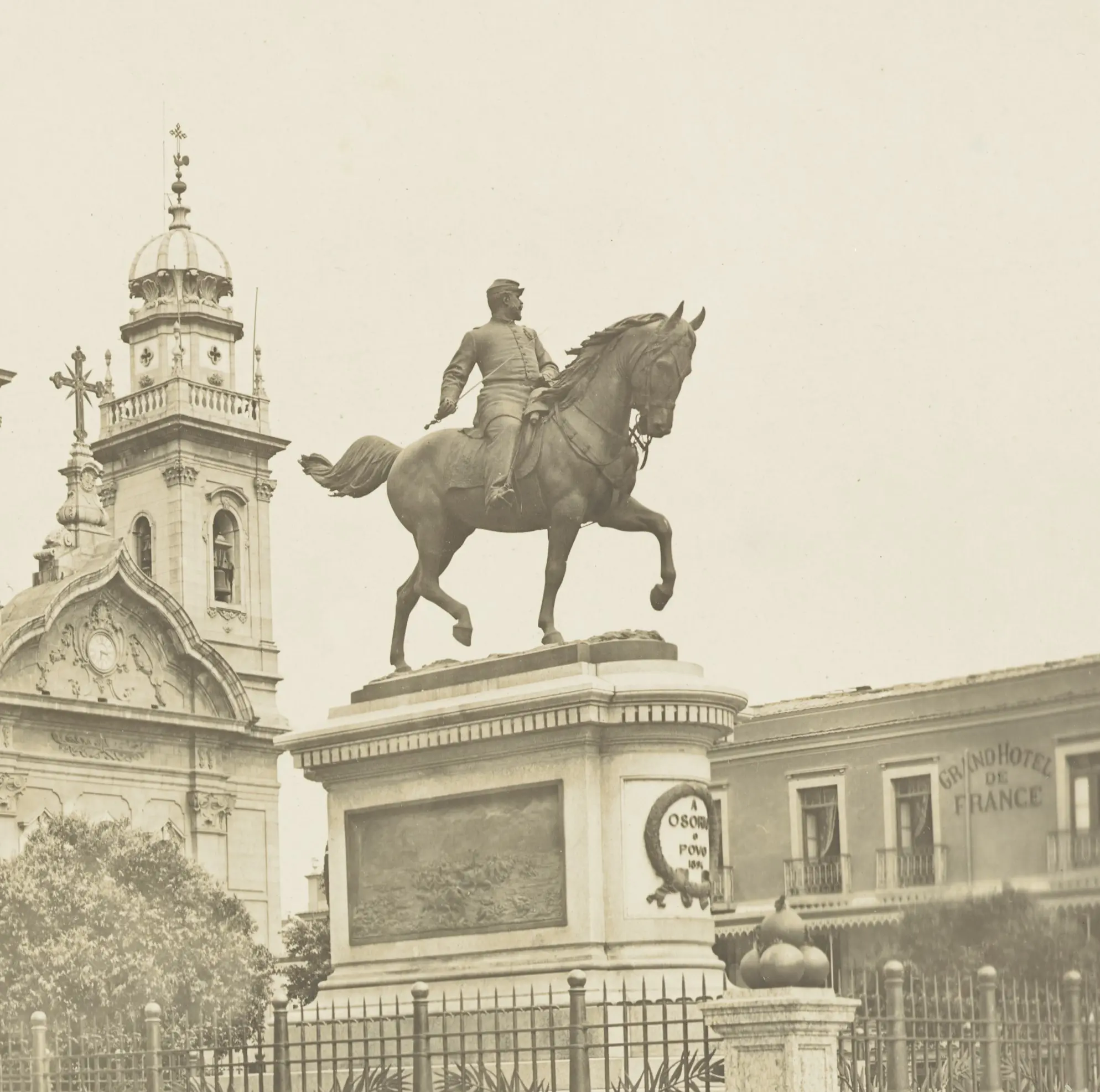 Estátua equestre de general em pedestal, torre de igreja à esquerda e edifício à direita.