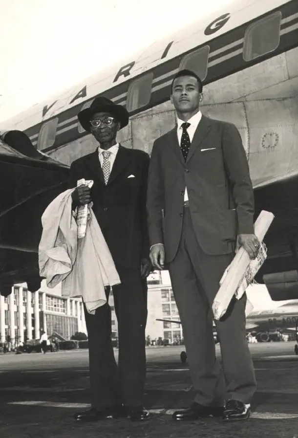 Dois homens em frente a um avião, um mais velho com jornal dobrado e outro jovem com jornal aberto.
