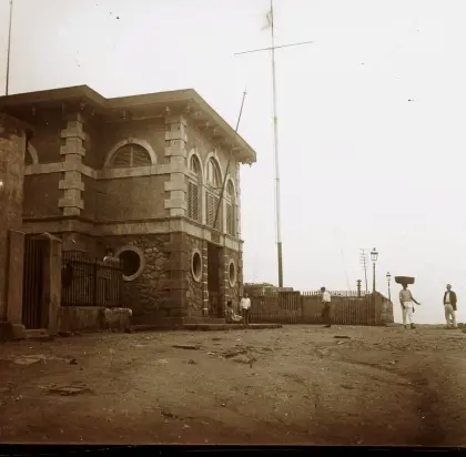 Edifício antigo com arquitetura histórica, pessoas caminhando e antena de rádio ao fundo.
