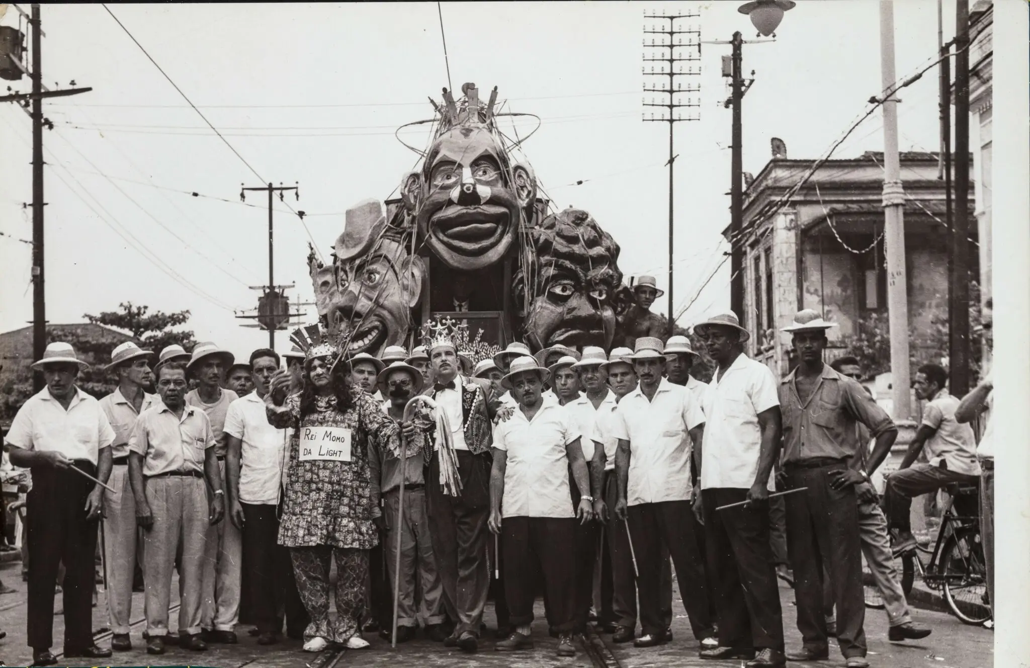 Grupo de pessoas em frente a estrutura monumental, com figura central vestida coloridamente segurando cartaz.