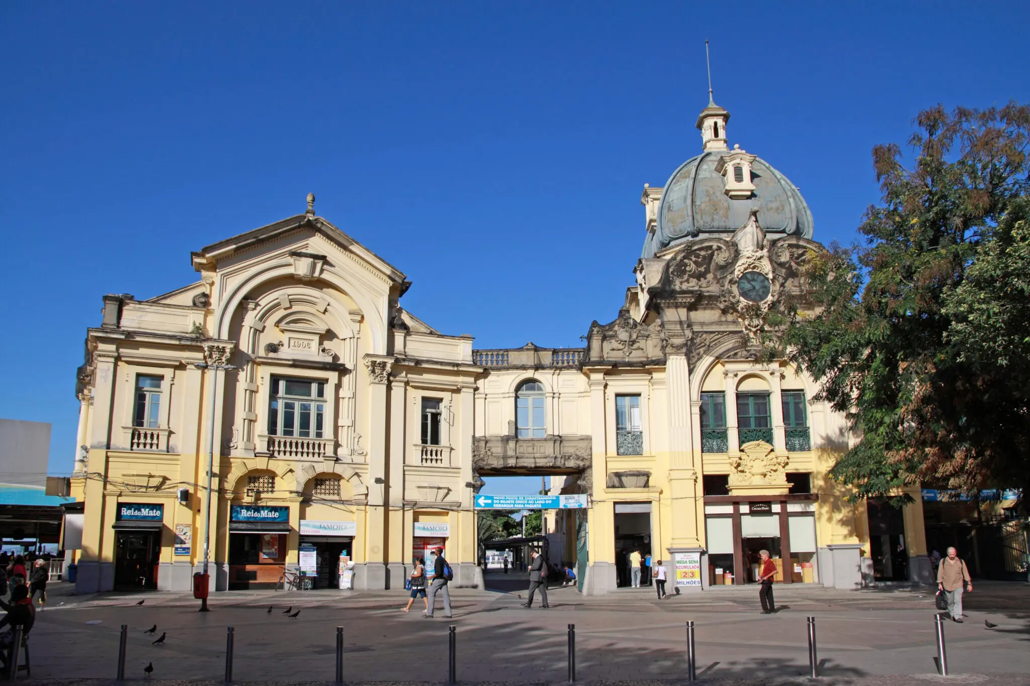 Praça histórica com edifício amarelo e cúpula azul, pessoas caminhando e árvores à direita.