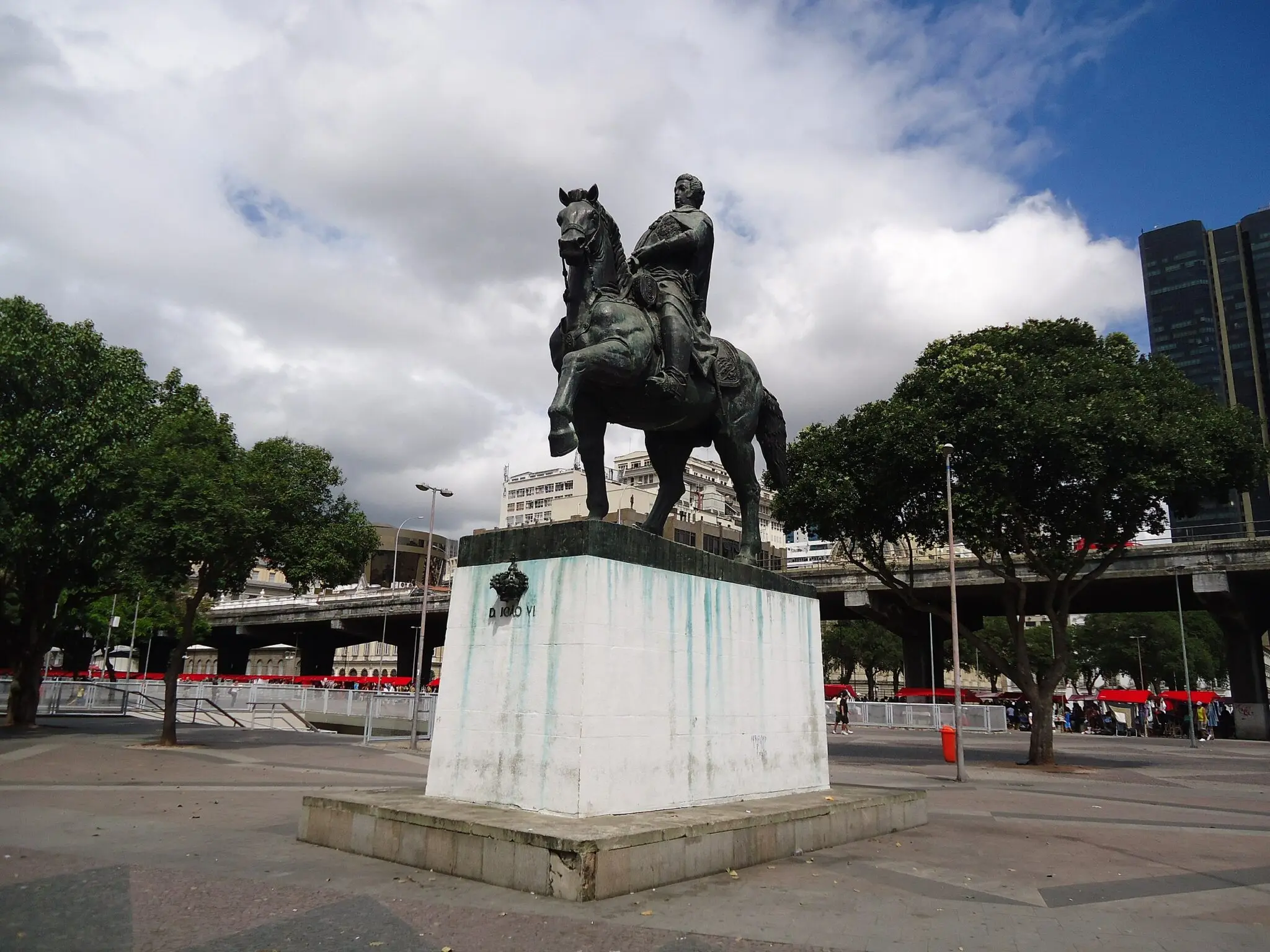 Estátua equestre em praça pública, com árvores e edifícios ao fundo.