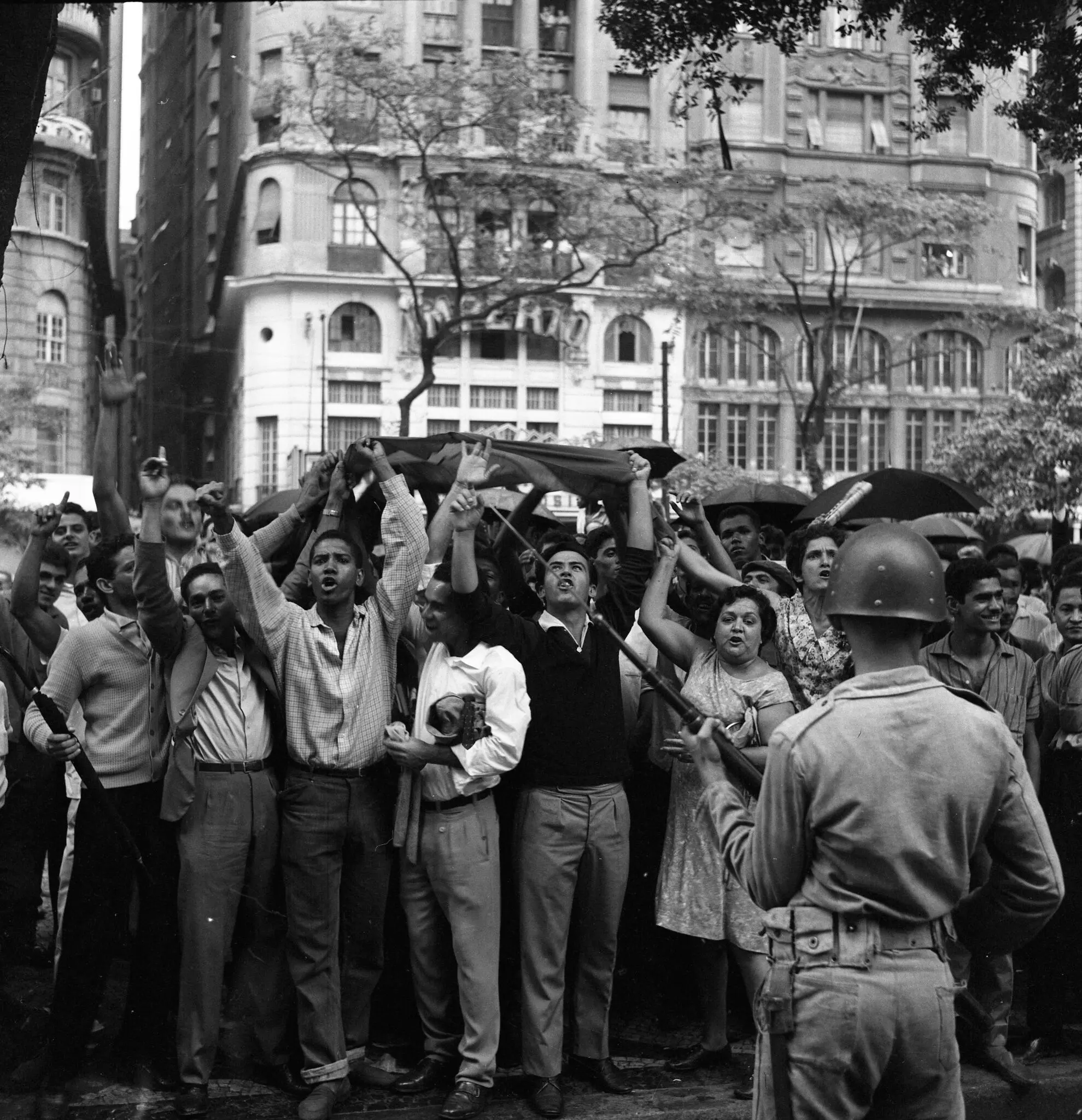 Manifestação pública com pessoas levantando braços e policial apontando arma.