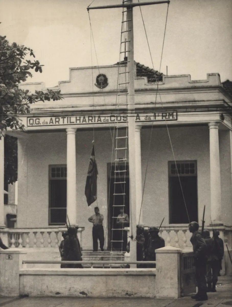 Edifício antigo com inscrição, colunas e varanda, árvore à esquerda, homens com bandeira e lanças à direita.