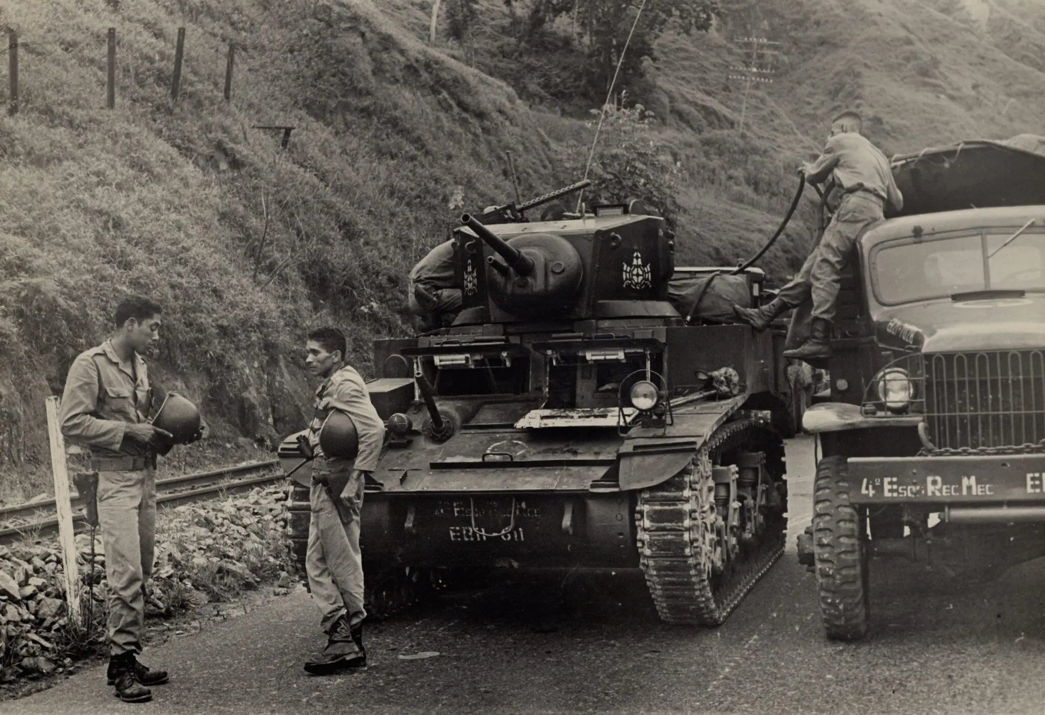 Cena militar com tanque e veículos blindados em trilhos.