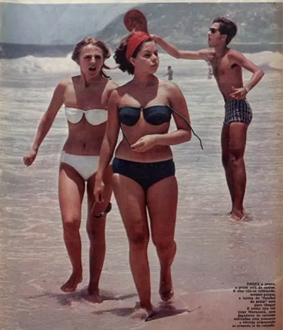 Três pessoas na praia: duas mulheres caminhando e um homem conversando.