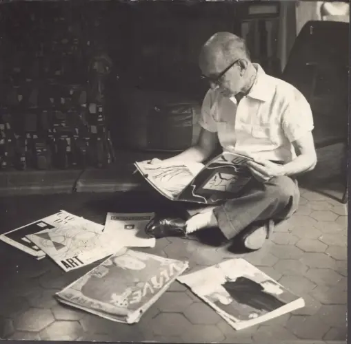 Homem lendo jornal no chão, óculos e camisa branca.