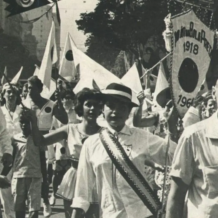 Procissão festiva em preto e branco, com pessoas vestidas festivamente, bandeiras e árvores no fundo.