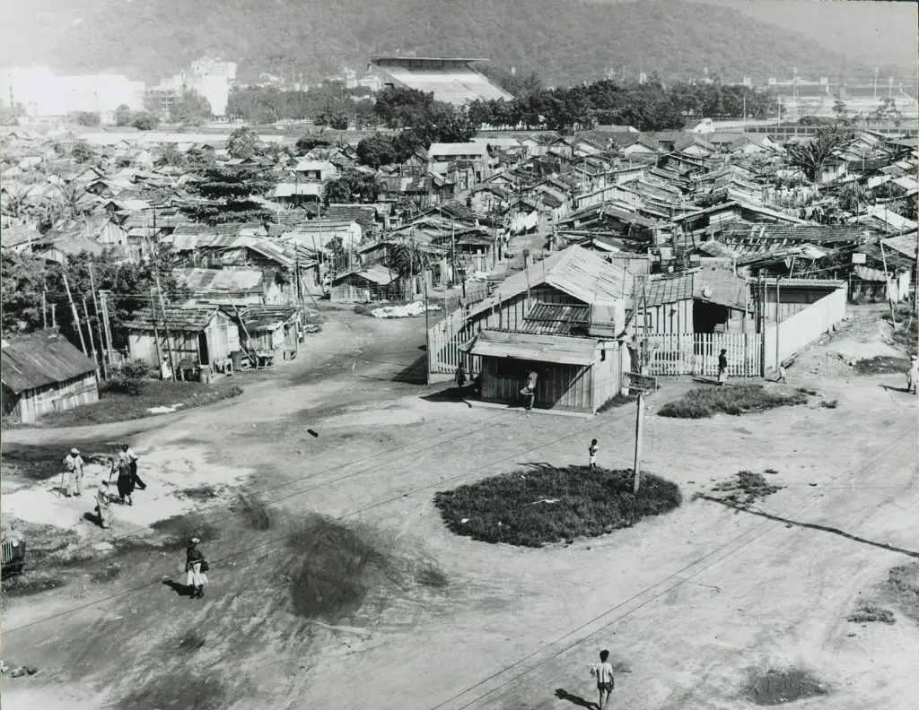 Fotografia preto e branco de favela com casinhas de madeira e edifício inclinado.