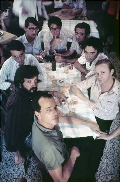 Grupo de pessoas reunidos em torno de mesa com guardanapo xadrez, bebidas e conversa social.