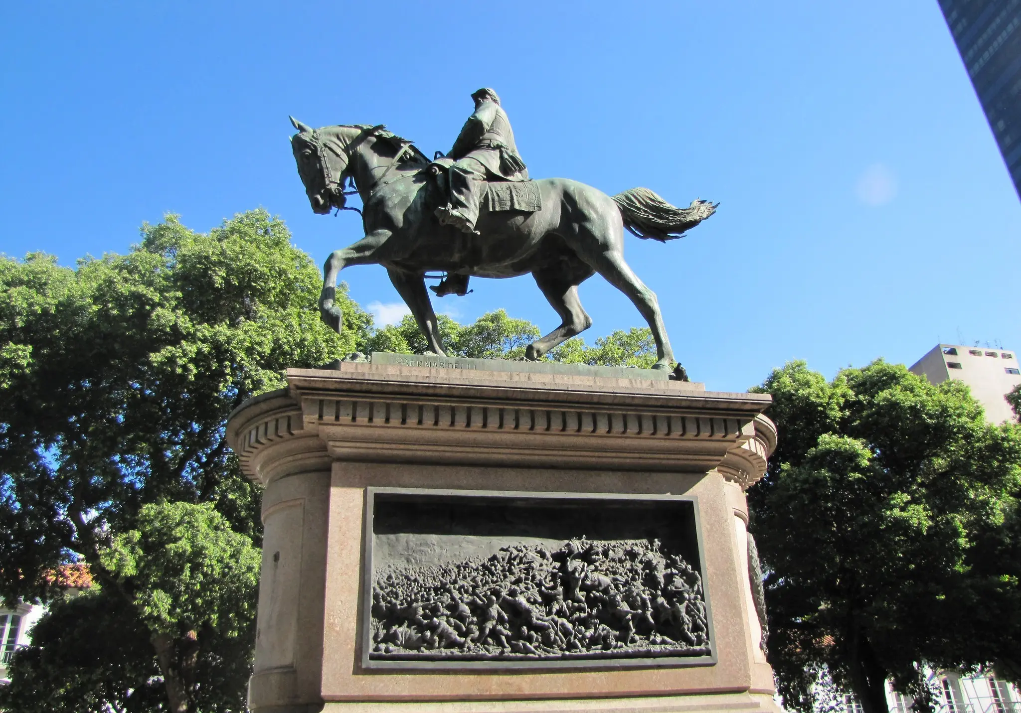 Estátua de general em cavalo em praça pública, com árvores e edifícios ao fundo.