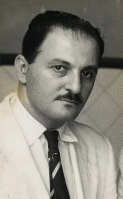 Homem sentado, blazer claro e camisa branca com gravata preta e branca.