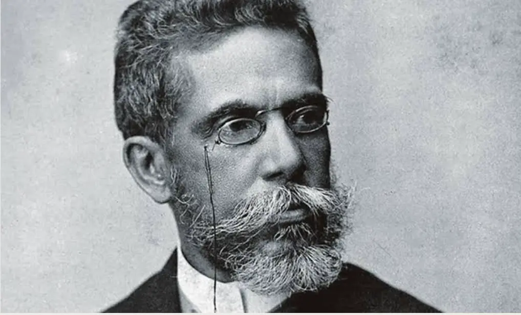 Homem adulto de perfil, terno clássico, cabelos grisalhos, barba densa, fundo claro.