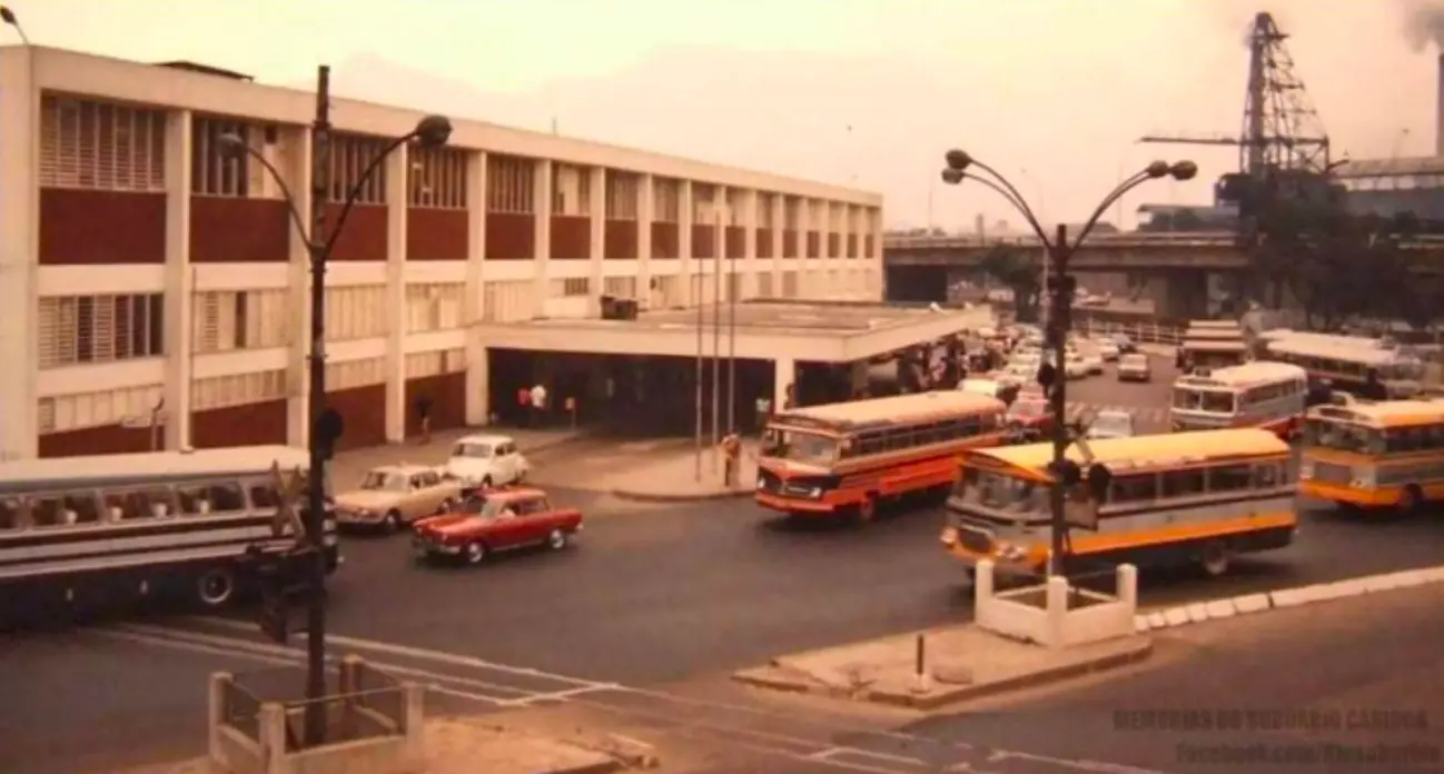 Rodoviária com edifício branco e detalhes vermelhos, pessoas caminhando, ônibus e carros coloridos.