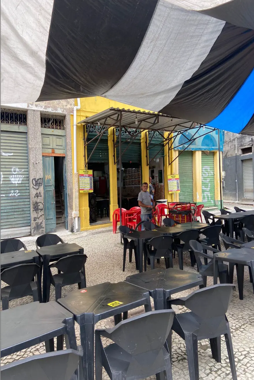 Bar em rua com edifícios coloridos, mesas pretas e pessoa caminhando.