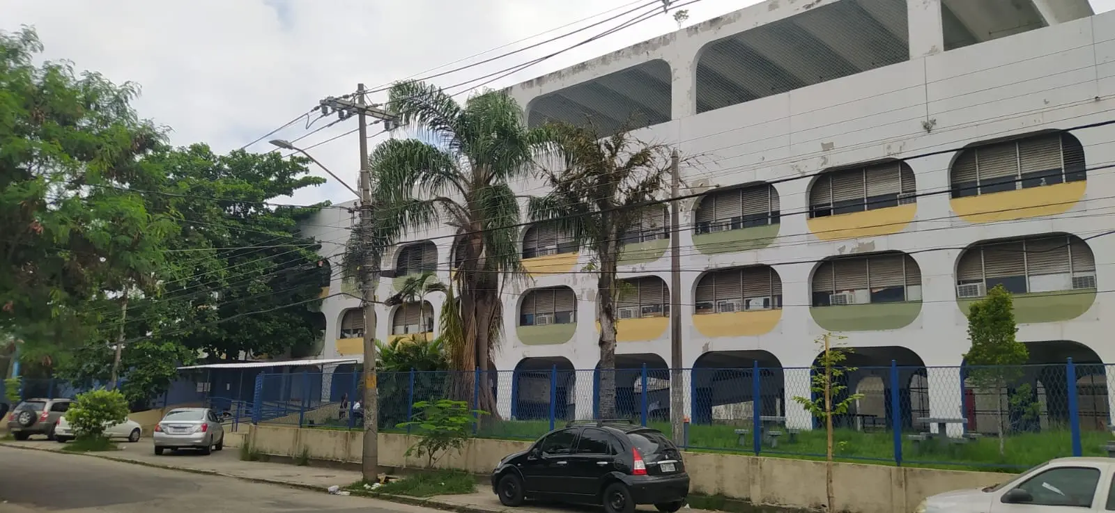Prédio escolar em bairro residencial com árvores, palmeiras e céu parcialmente nublado.