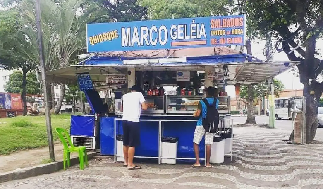 Quiosque de salgados em ambiente urbano, com árvores e pessoas ao redor.
