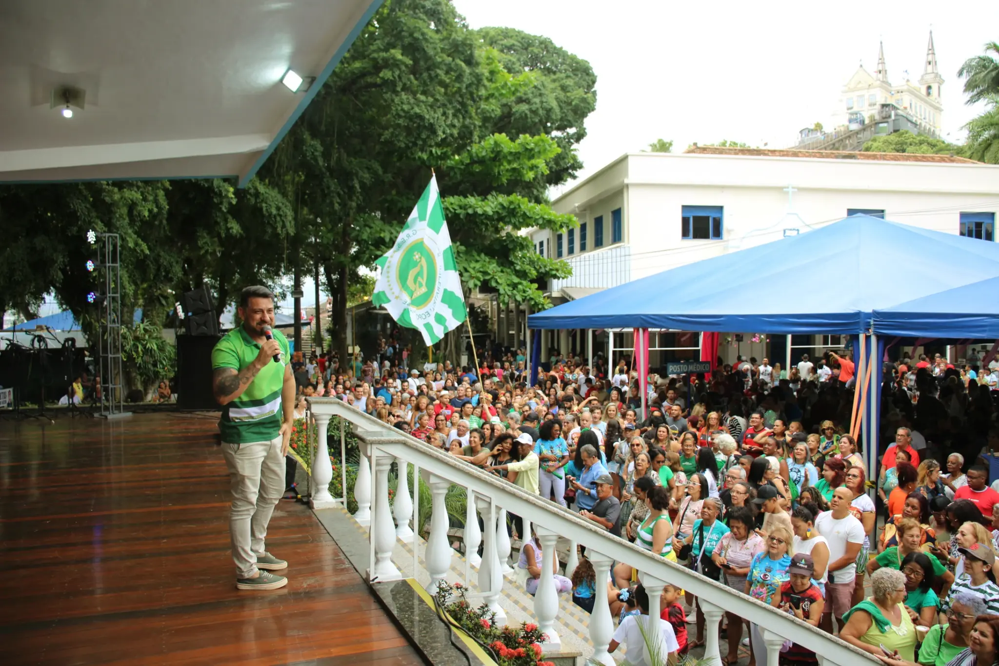 Pessoas em palco com microfone, estruturas metálicas e plantas verdes ao fundo.