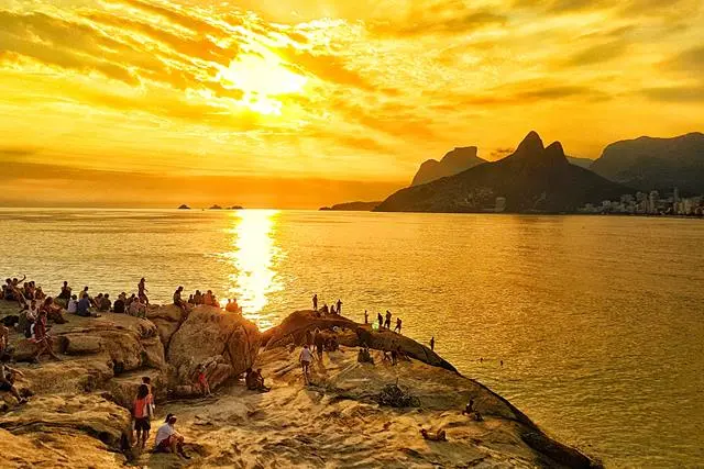Pôr-do-sol em praia com rochas, sol refletindo luz dourada e montanhas.