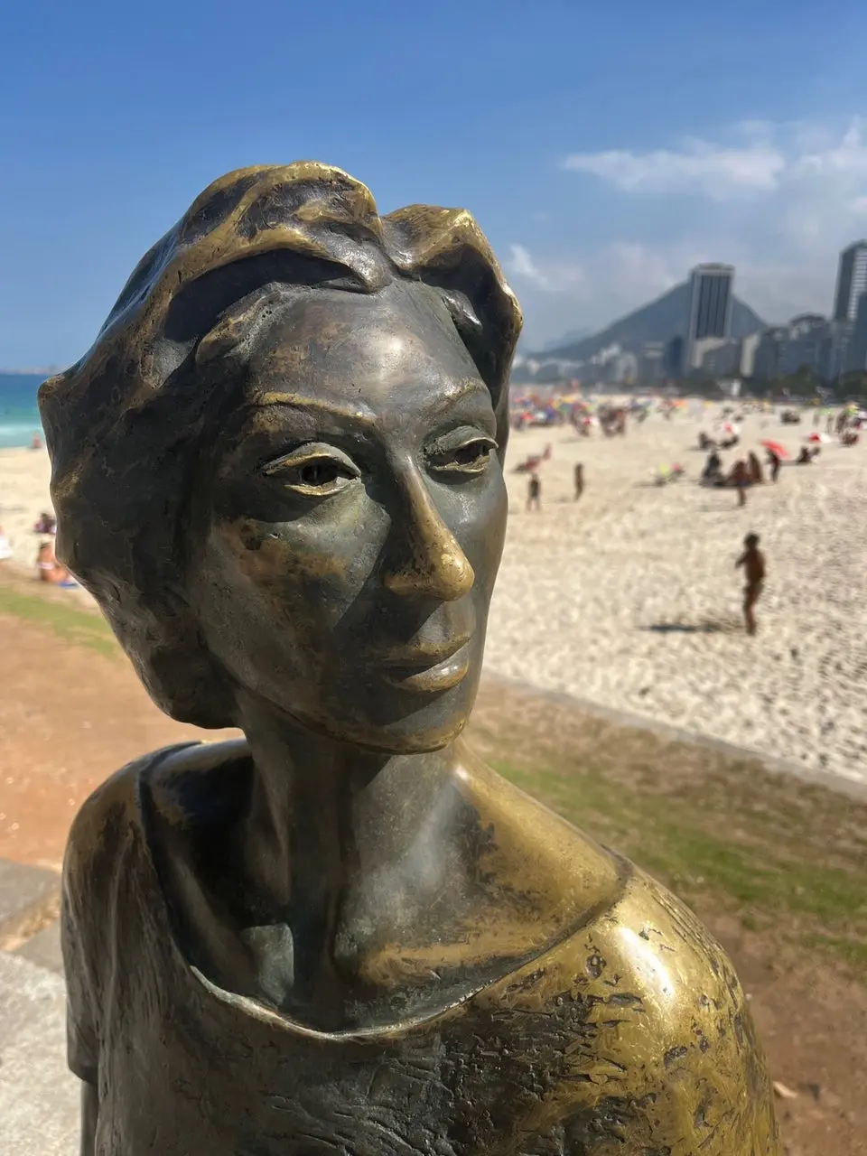 Estátua de bronze de mulher em praia com mar e montanha ao fundo.