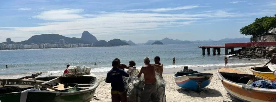 Praia com pessoas e barcos, mar calmo e montanhas verdes.