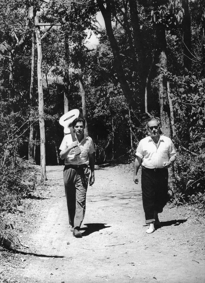 Dois homens caminham juntos em um caminho de terra, um segurando uma guitarra acústica.