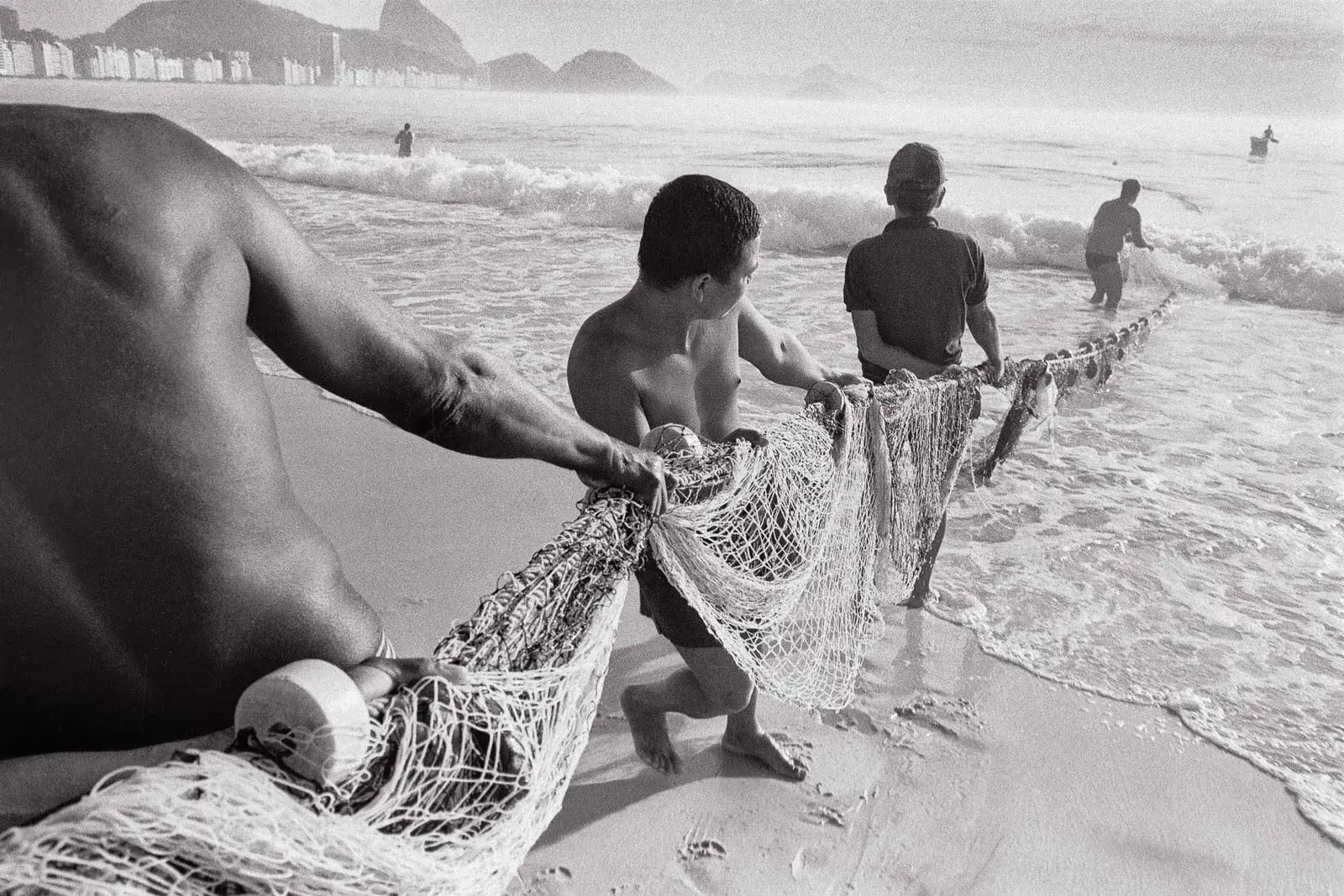 Colônia de pescadores em praia, com homem segurando rede e outros arrastando pela areia.