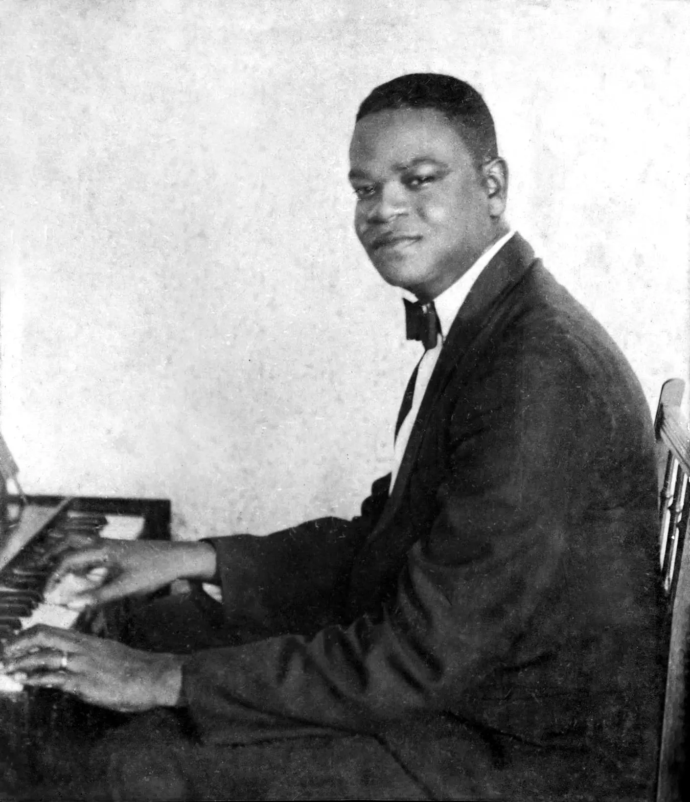 Homem em terno preto com gravata borboleta, tocando piano em foto preto e branco.