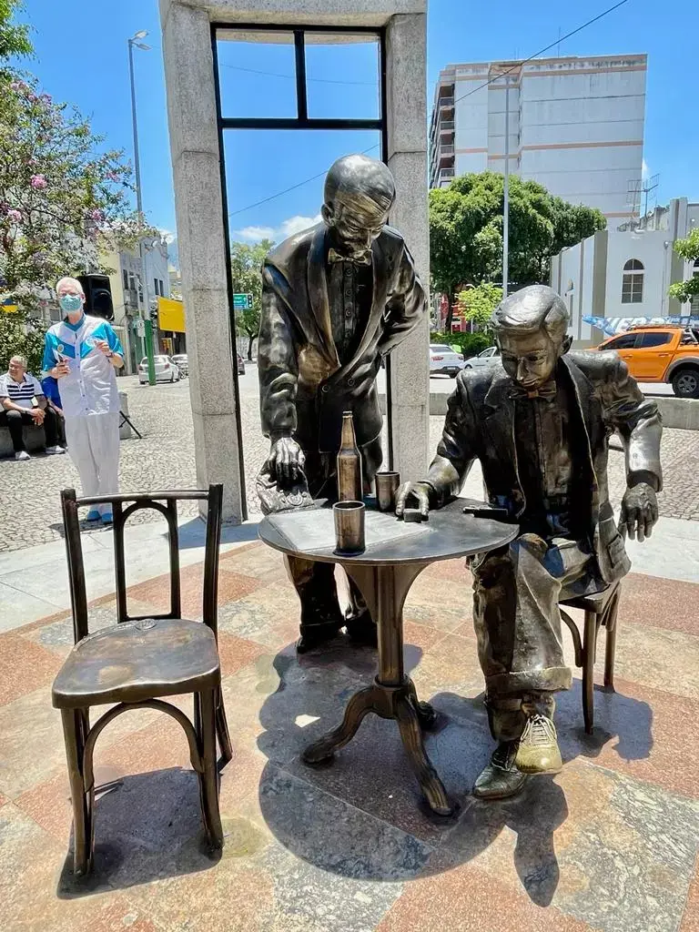 Estátua de bronze homenageando Noel Rosa, com dois homens interagindo em torno de uma mesa com cerveja.