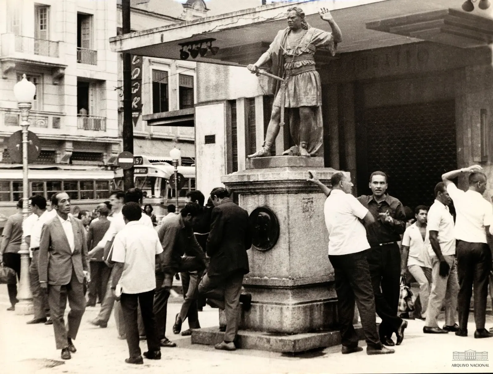 Praça preto e branco com estátua monumental e pessoas caminhando.