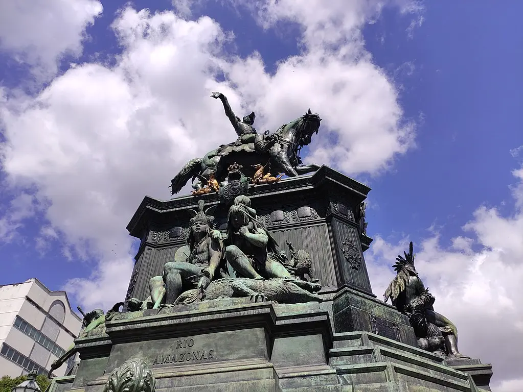 Estátua monumental em Tiradentes Square, figura a cavalo segurando dístico, base quadrada com detalhes ornamentais, figuras humanas e animais, céu azul parcialmente nublado.