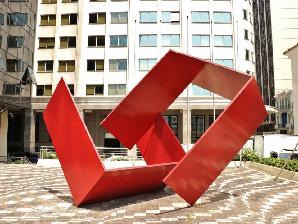 Escultura geométrica vermelha em frente a edifício moderno.