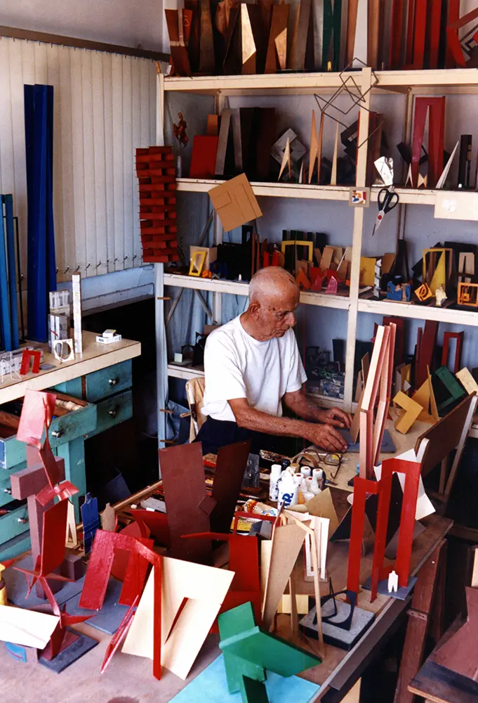 Homem trabalhando em estúdio de escultura com objetos artesanais coloridos.