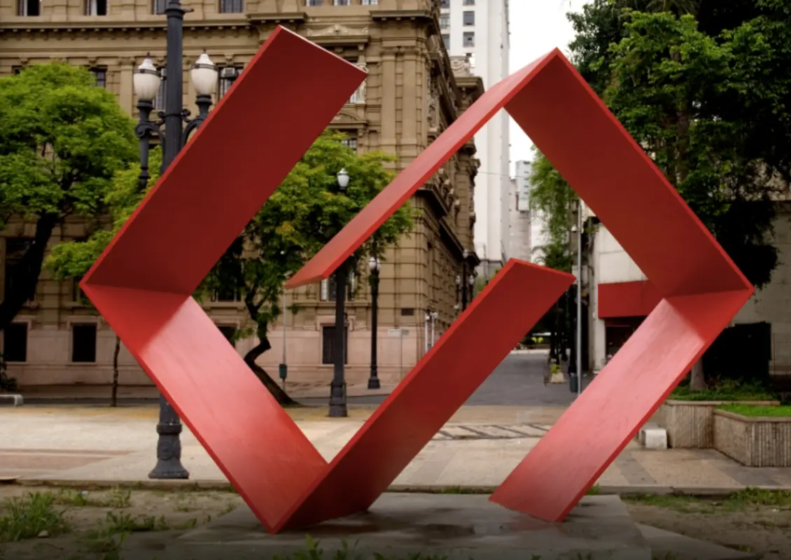 Escultura urbana com cruz invertida vermelha, edifícios históricos e modernos ao fundo.