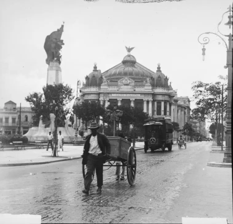 Praça com monumento, teatro, estátua monumental, pessoa caminhando, carrinho, edifício com cúpulas, lâmpada de rua e carruagem de cavalos.