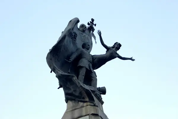 Estátua em preto e branco com figuras voando e levantadas contra céu claro.