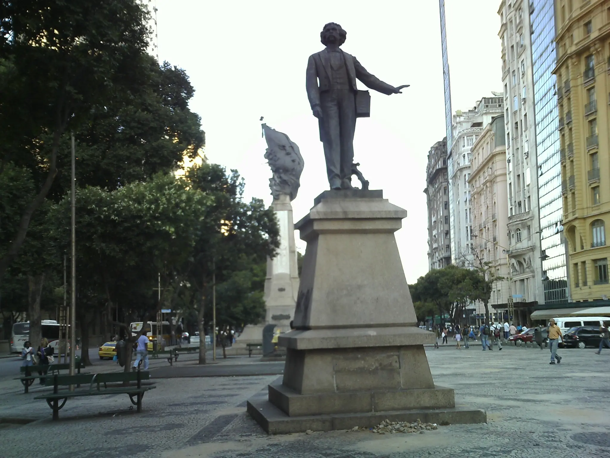Estátua de bronze em praça pública com árvores e edifícios ao fundo.