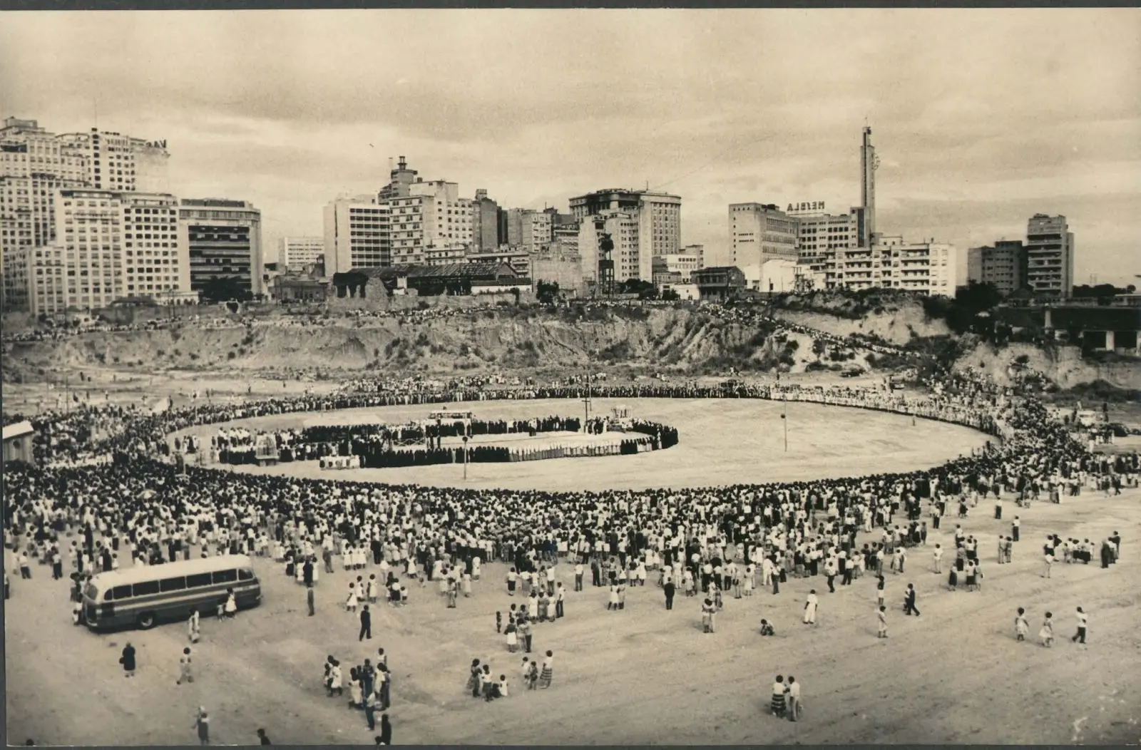 Evento em praça com pessoas em círculo e edifícios modernos ao fundo.