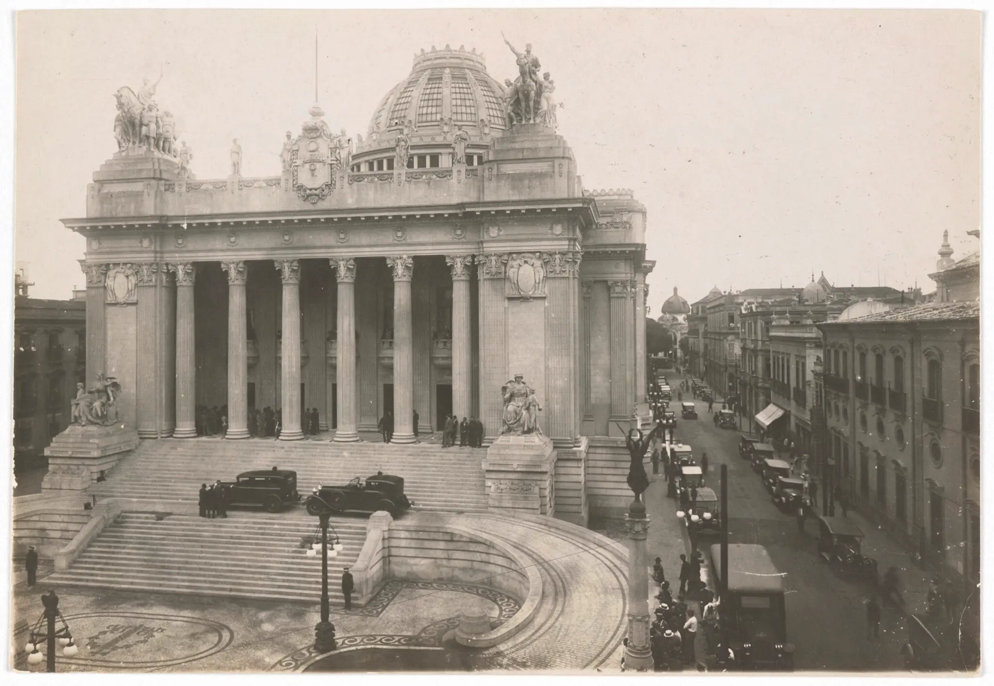 Palácio monumental com escadaria, pessoas caminhando, estátuas e colunas.