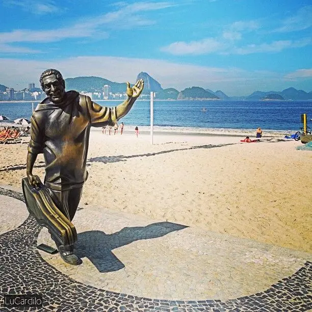Estátua de bronze em praia com pessoas caminhando e montanhas ao fundo.