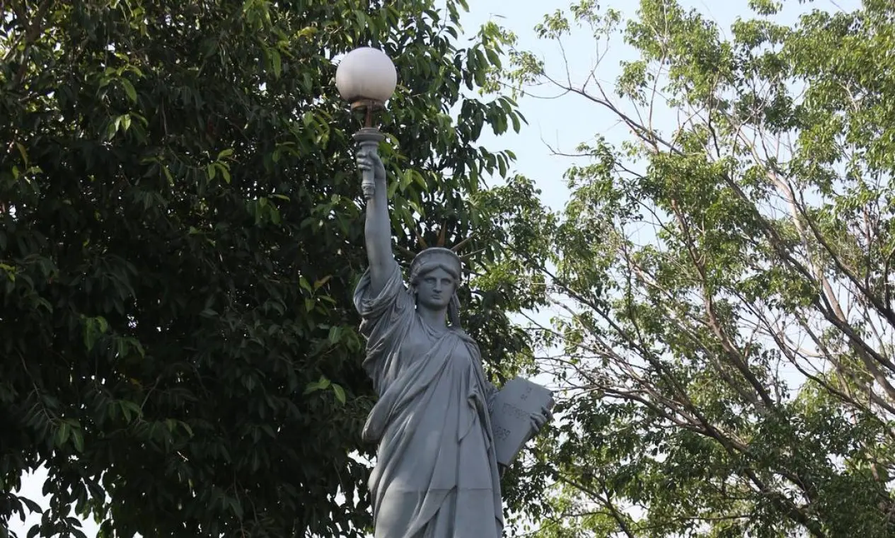 Estátua da Liberdade de Vila Kennedy, cinza, lanterna na mão, livro aberto à frente, árvores verdes ao fundo.