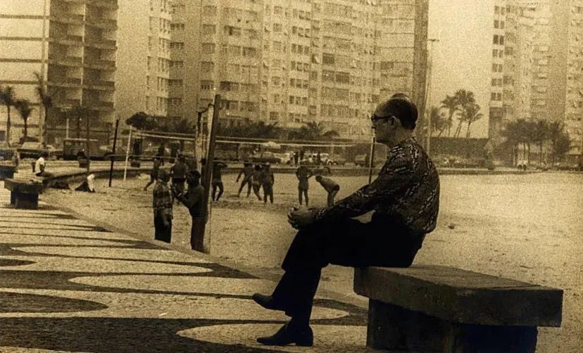 Homem sentado em banco, pátio com futebol, edifícios altos ao fundo.