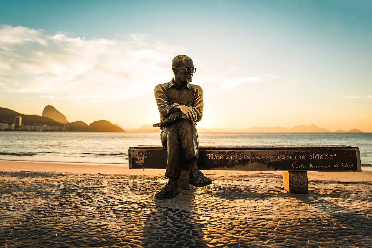 Estátua de bronze de Carlos Drummond de Andrade em frente à praia, com mar e montanhas ao fundo.