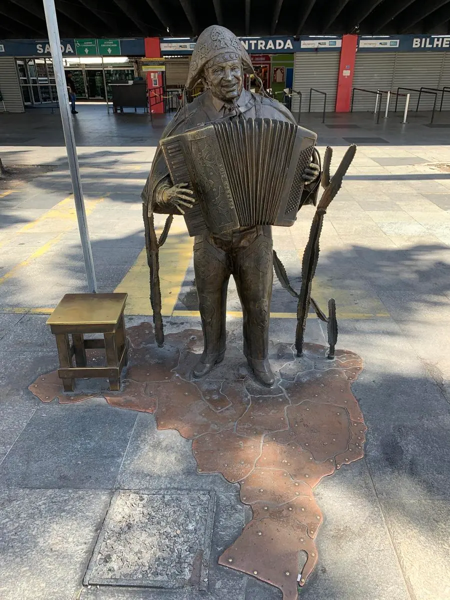 Estátua de bronze de homem tocando acordeão, com base decorativa em forma de mapa.