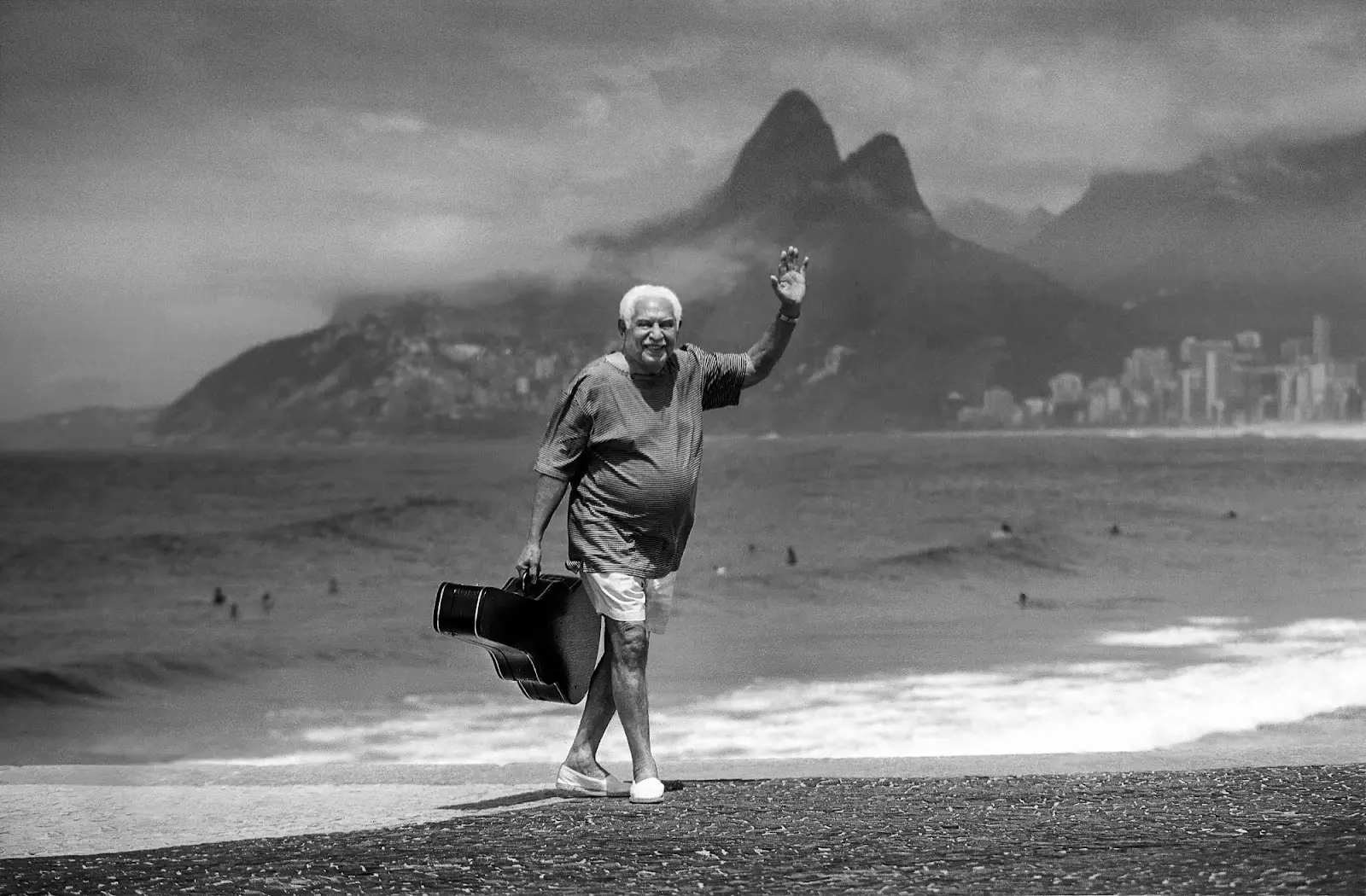 Homem sorridente caminhando em praia com montanhas e cidade ao fundo.