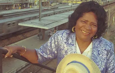 Mulher sorridente em blazer azul floral, chapéu de palha amarelo, perto de balaustrada metálica, plataforma de trem ao fundo.