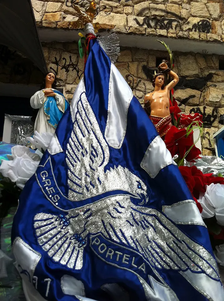 Decoração festiva com bandeira azul e branca, estátuas feminina e masculina segurando objetos dourados.