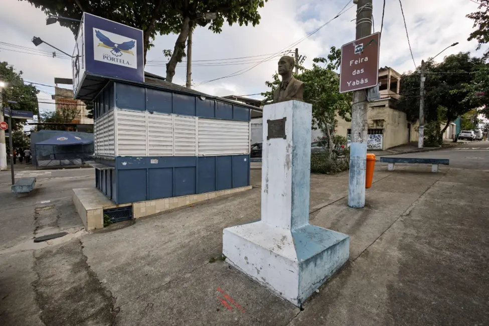 Praça urbana com edifício azul, águia, monumento e sinal "Feira das Yabás".