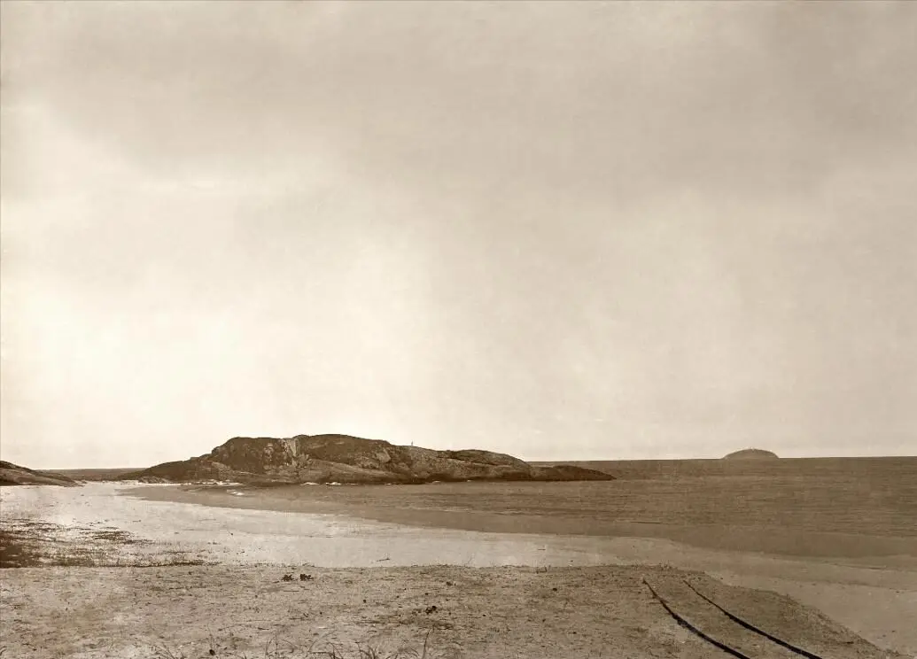 Paisagem costeira em sepia com praia arenosa, formação rochosa e céu cinzento.