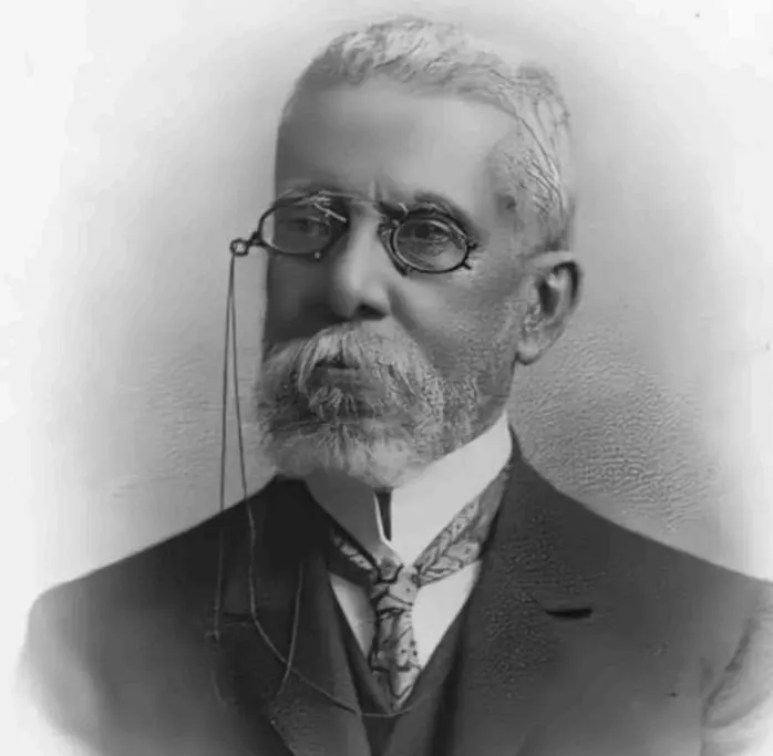 Homem adulto em terno clássico, barba grisalha, óculos com cordão, retrato preto e branco.